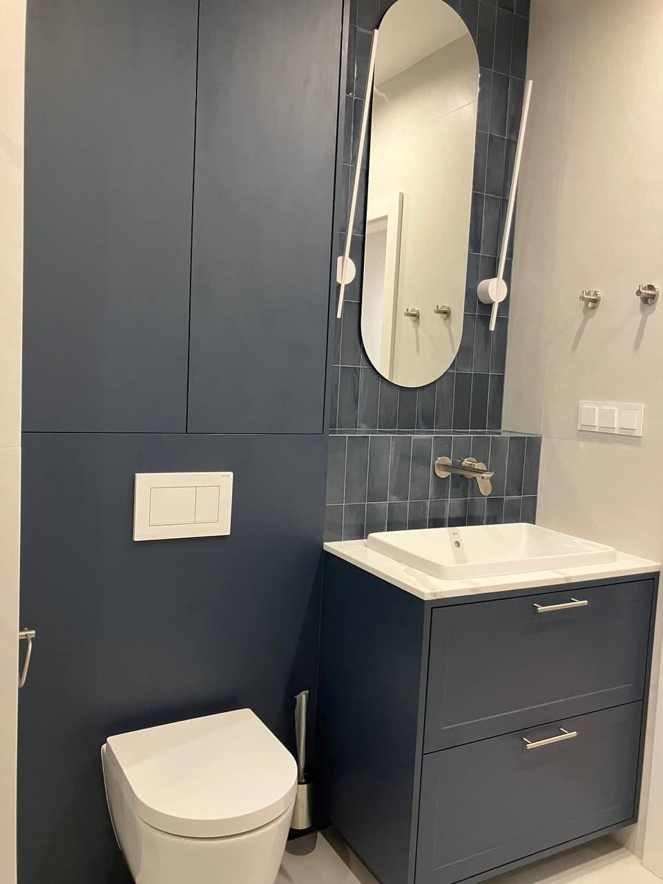 Granatowa zabudowa łazienkowa z szafką podumywalkową, zabudową stelaża WC i owalnym lustrem